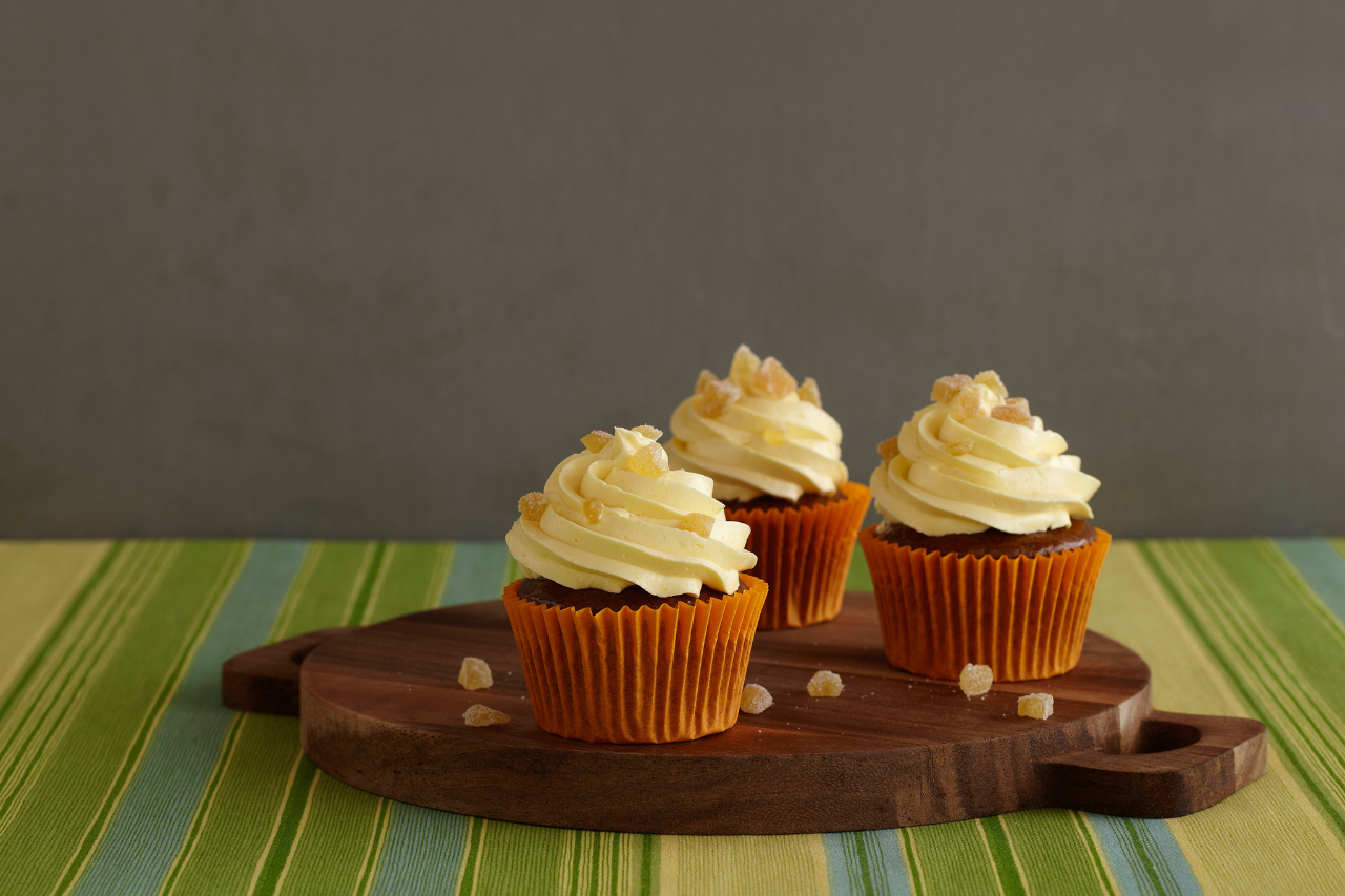 A wooden cutting board with three iced cupcakes topped with candied ginger