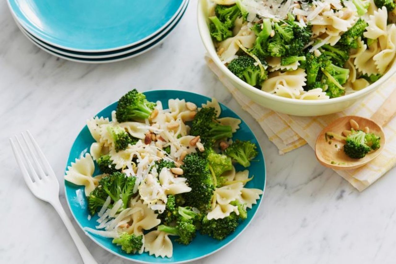 A blue plate with broccoli, bow tie pasta and cheese with a larger serving dish nearby