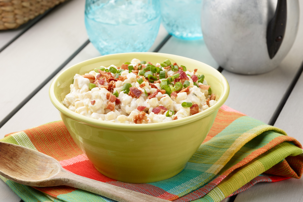 A bowl with creamed corn topped with fresh herbs and bacon sits on a picnic table
