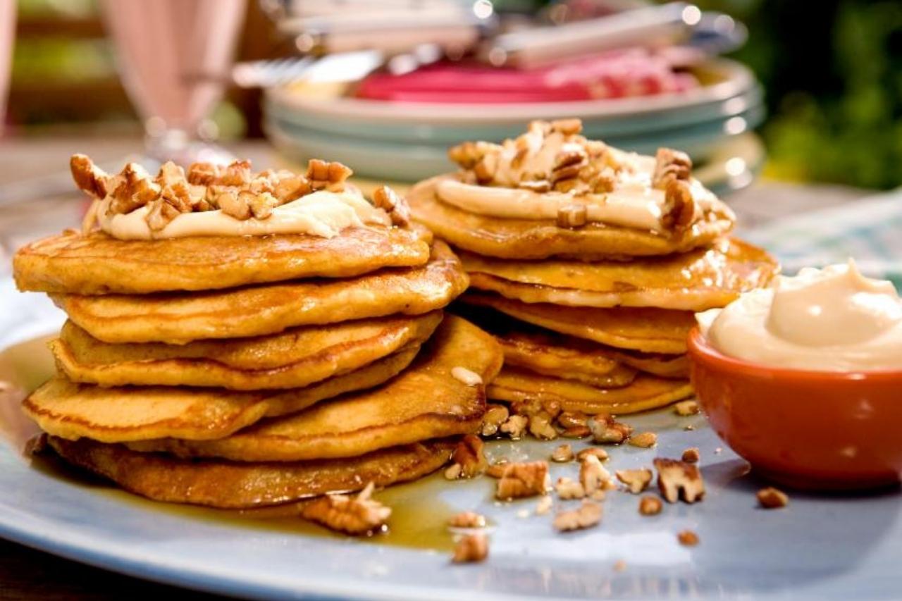 A large stack of fluffy carrot-cake flavoured pancakes with a cream cheese topping and chopped pecans