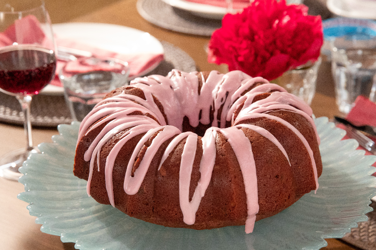 A double chocolate bundt cake with drizzled pink icing on a plate near a display of roses