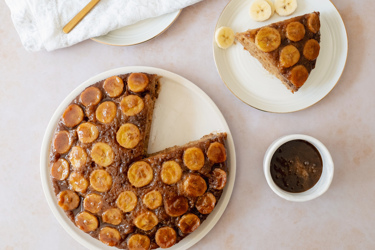 Overhead shot of banana upside down cake, one slice cut out and a small bowl of toffee sauce nearby