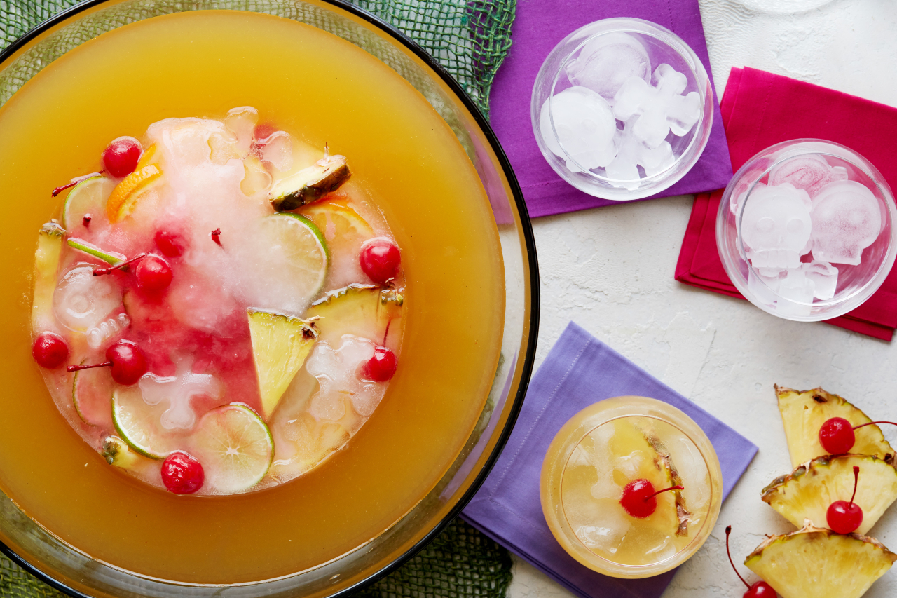 A bowl of spiced rum punch topped with fresh fruit, with a glass of punch and a glass of ice on the side