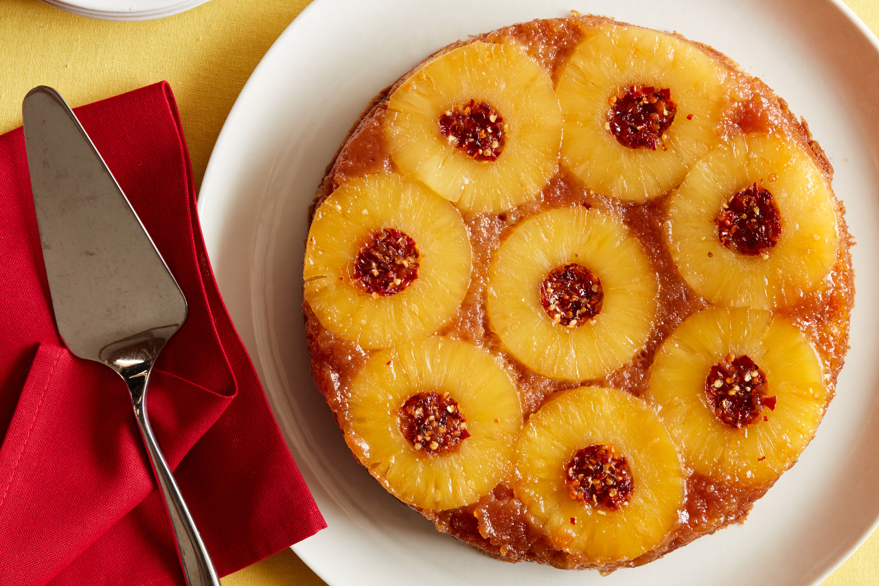 Pineapple upside down cake topped with pineapple rings and candied chile peppers served on a white cake plate