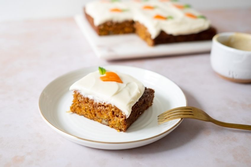 A slice of square carrot cake with cream cheese frosting on a white round plate. A fork is placed on the plate. You can see the rest of the cake and a cup in the background.