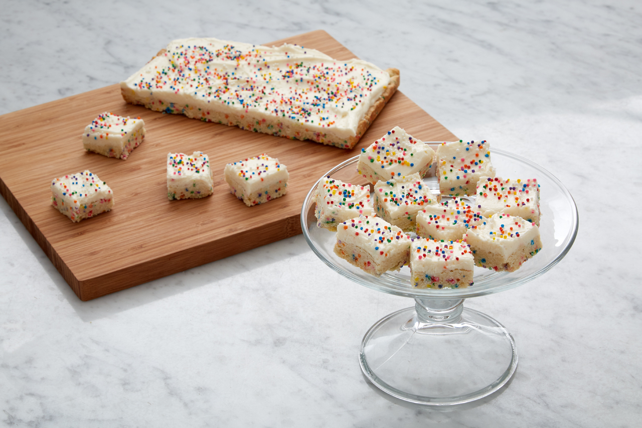 A dessert tray with confetti frosted sugar cookie squares with more on a wooden cutting board