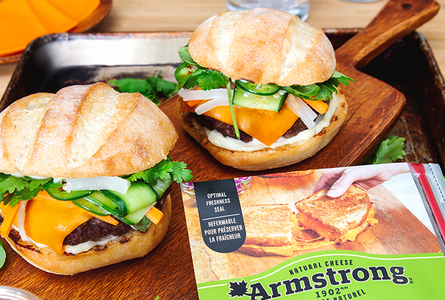 Two beef burgers with a slice of Armstrong Medium Cheddar Cheese, fresh herbs and quick pickles sit on a wooden board, a package of Armstong Cheese beside them