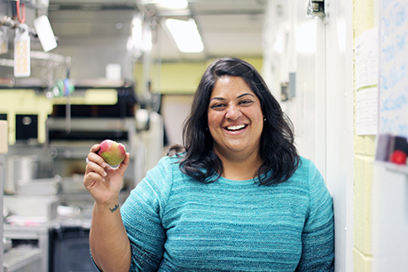 Joshna Maharaj standing in a professional kitchen, holding an apple and smiling
