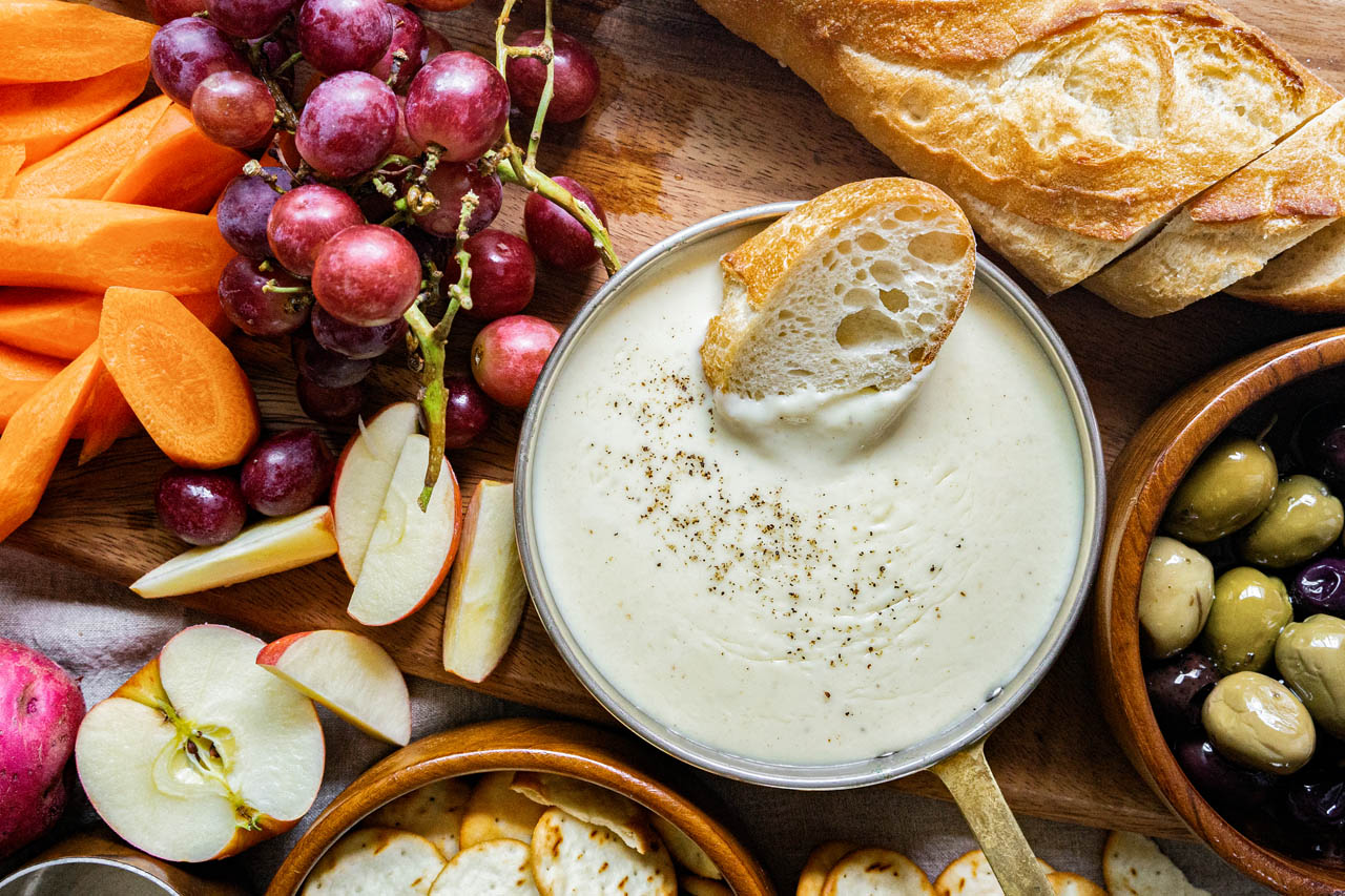 cheese fondue board surrounded with bread, olives, grapes, carrots