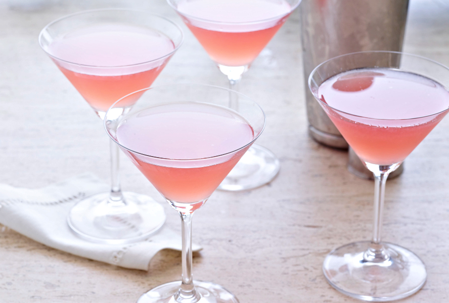 A cocktail shaker and four martini glasses filled with pink cosmo cocktails