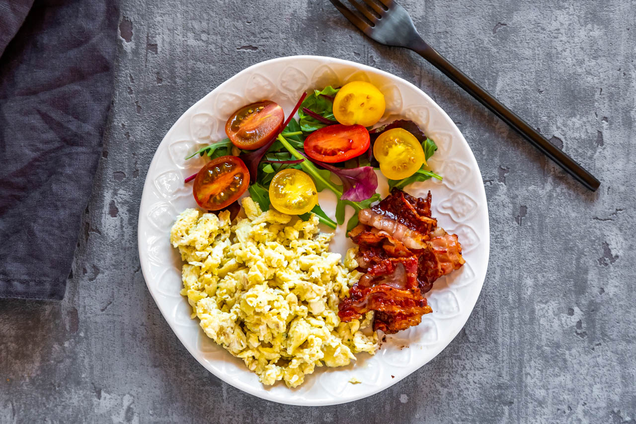A keto-approved plate of scrambled eggs, bacon and veggies on a grey tabletop