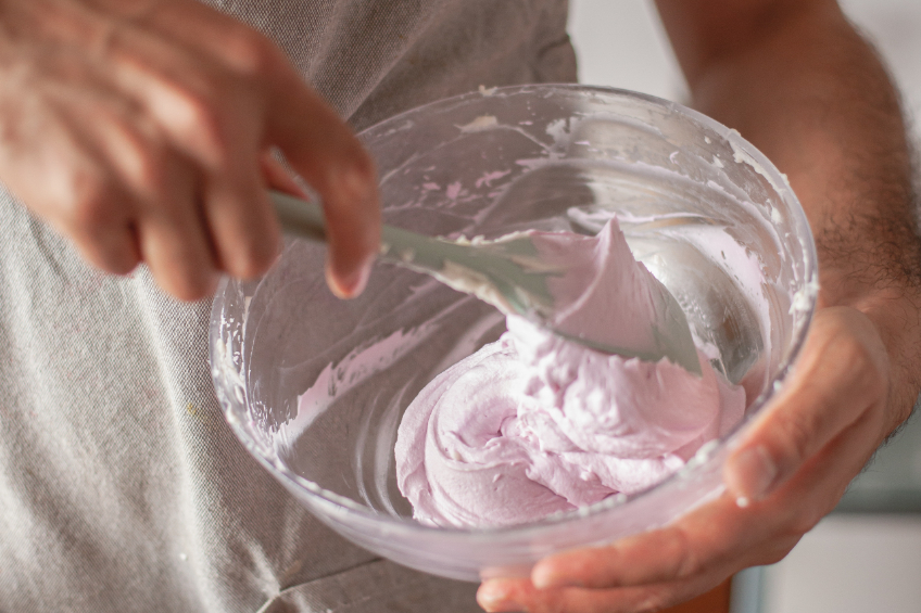 A hand mixes some pink buttercream in a glass mixing bowl