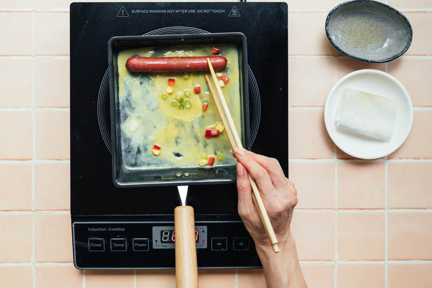 a hotdog placed over omelette mixture in a square pan, ready to be rolled