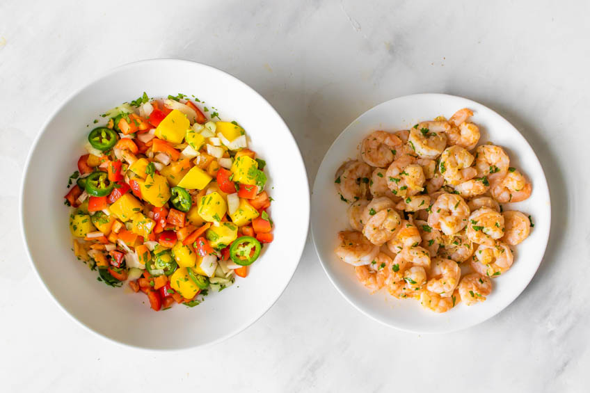 A bowl of spicy mango salsa beside a plate of cooked shrimp on a countertop