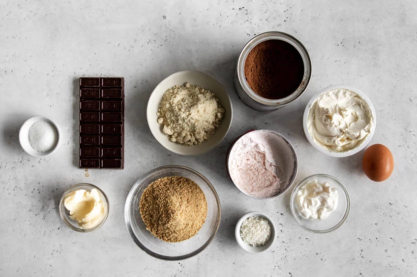 A flat lay of the ingredients needed for Nanaimo bar cheesecake: chocolate, graham crackers, eggs, cream, sugar, butter and cocoa powder