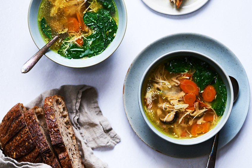 A bowl of bone broth, chicken and vegetable soup with shiitake mushrooms, carrots and baby spinach. A loaf of sliced bread is on the side.
