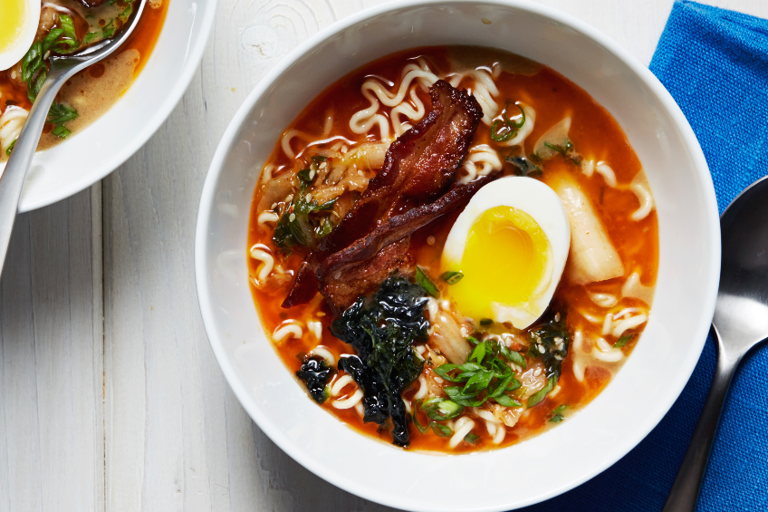 A white bowl filled with soup. It includes half of an egg, slices of bacon, ramen noodles, kimchi and nori