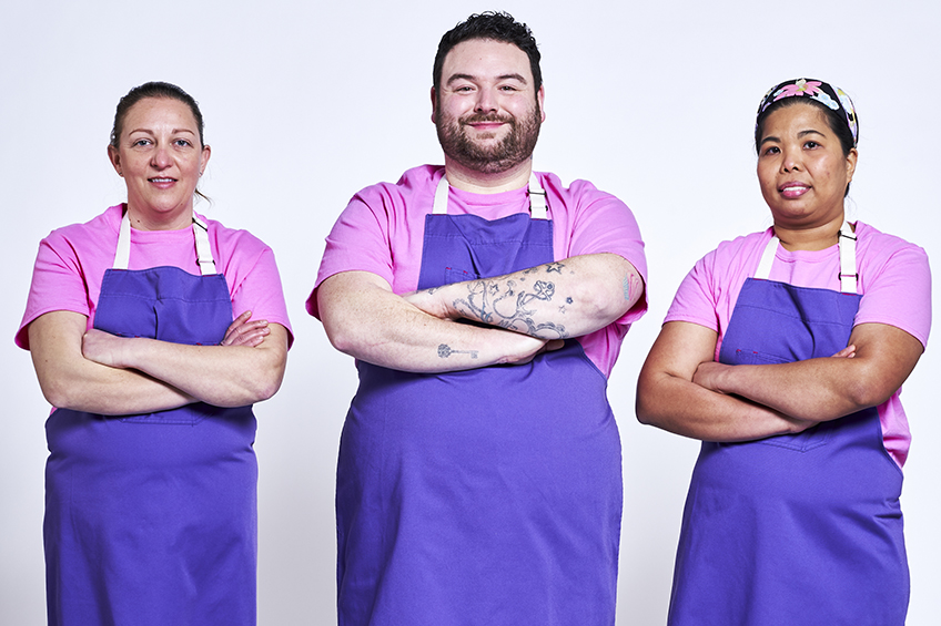 3 contestants looking at the camera and smiling wearing purple aprons