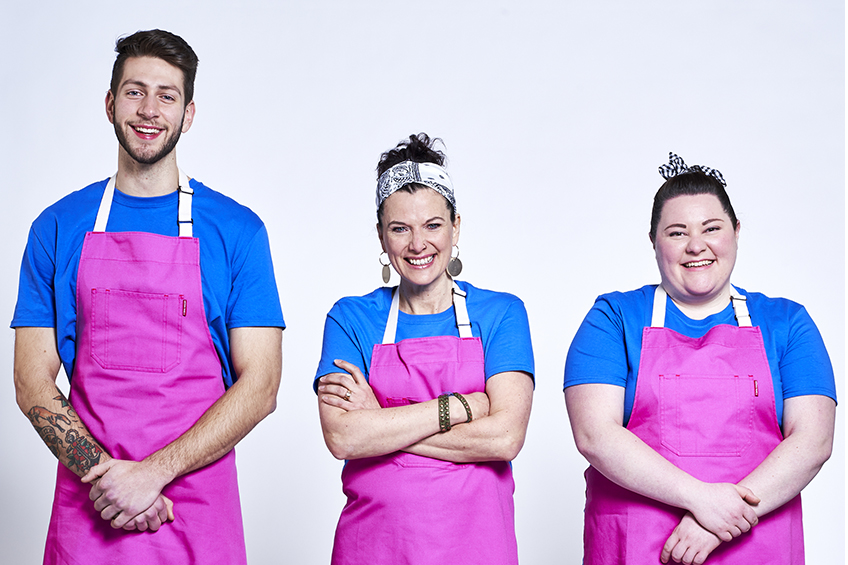 3 contestants looking at the camera and smiling wearing pink aprons