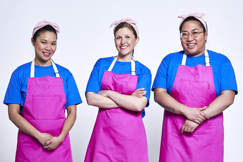 3 contestants looking at the camera and smiling wearing pink aprons