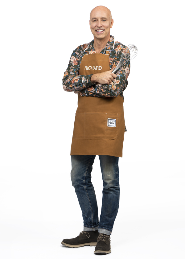 Richard Martemucci poses in a floral button down top and brown apron while holding a metal whisk