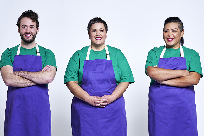 3 contestants looking at the camera and smiling wearing purple aprons