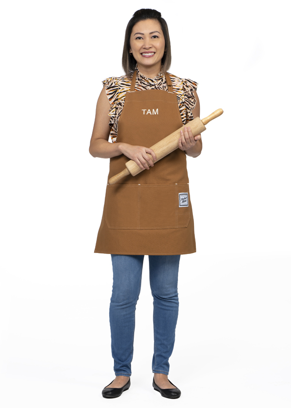 Tam Truong poses with a wooden rolling pin while wearing jeans and a brown kitchen apron