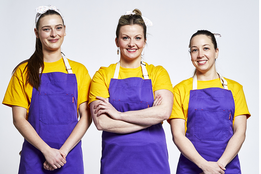 3 contestants in purple aprons smiling at the camera