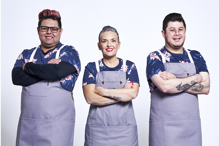3 contestants looking at the camera and smiling wearing grey aprons