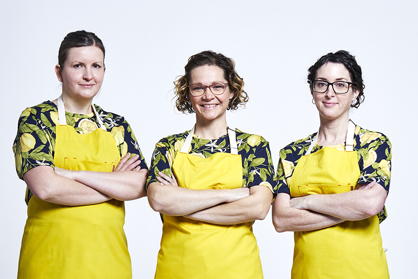3 contestants looking at the camera and smiling wearing yellow aprons