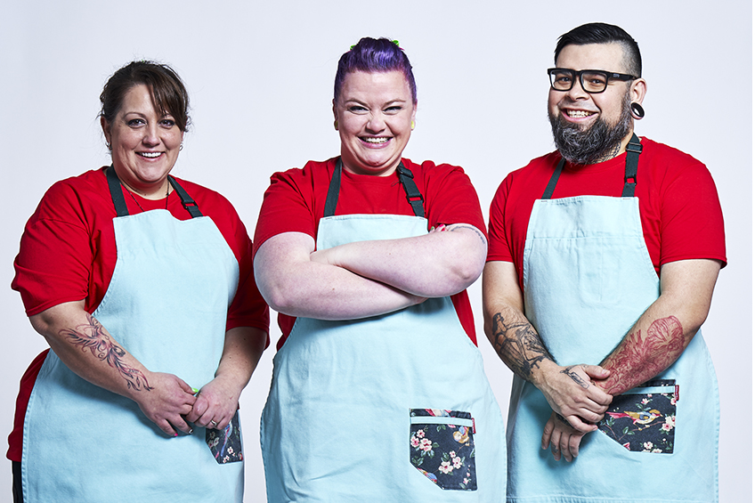 3 contestants looking at the camera and smiling wearing light blue aprons