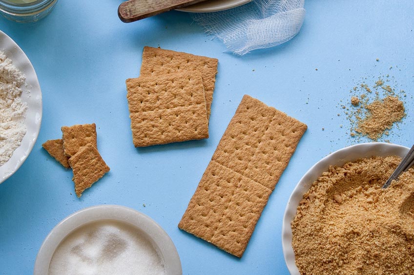 Graham crackers scattered on a blue countertop with bowls of sugar and graham cracker crumbs surrounding