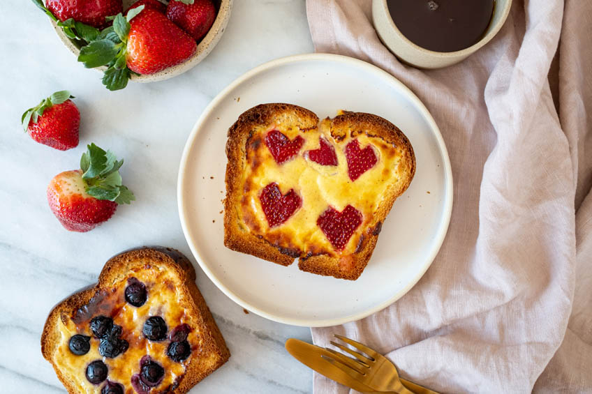 Toasted bread with custard in the centre and strawberry hearts on a white plate with a blueberry topped slice on the side