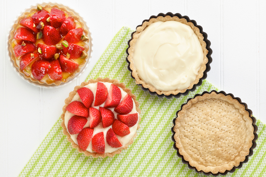 A glazed strawberry tart, an incomplete tart with pastry cream and strawberries, another incomplete tart with pastry cream, and an empty tart shell