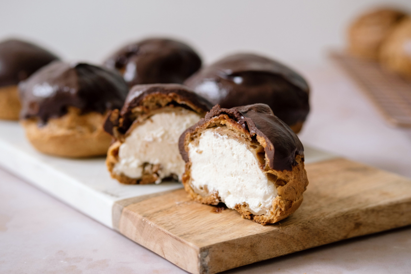 Cream puff with a mascarpone filling and chocolate ganache topping, cut in half, along with whole puffs on a wooden serving tray