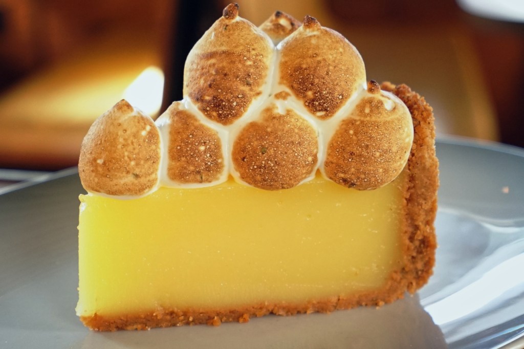 A plated slice of lemon meringue pie with bright yellow lemon curd and golden meringue in a graham cracker crust.
