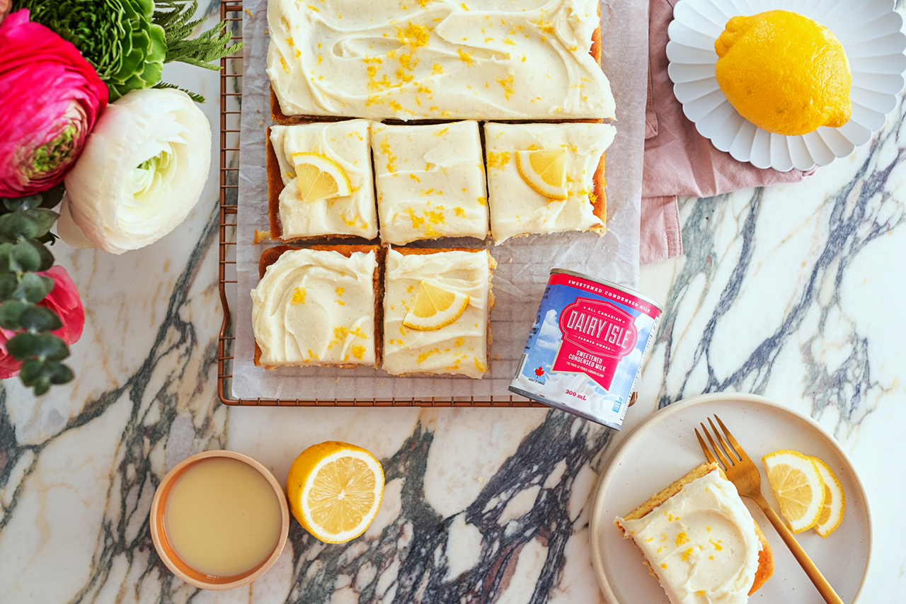 Overhead shot of a lemon sheet cake with lusciour sour cream icing with lemons, sweetened condensed milk and a perfectly cut slice laid out on a plate