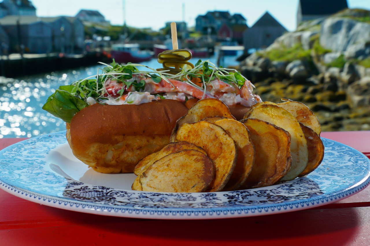 Lobster roll with a side of potato chips served on a blue plate with a seaside cove in the background