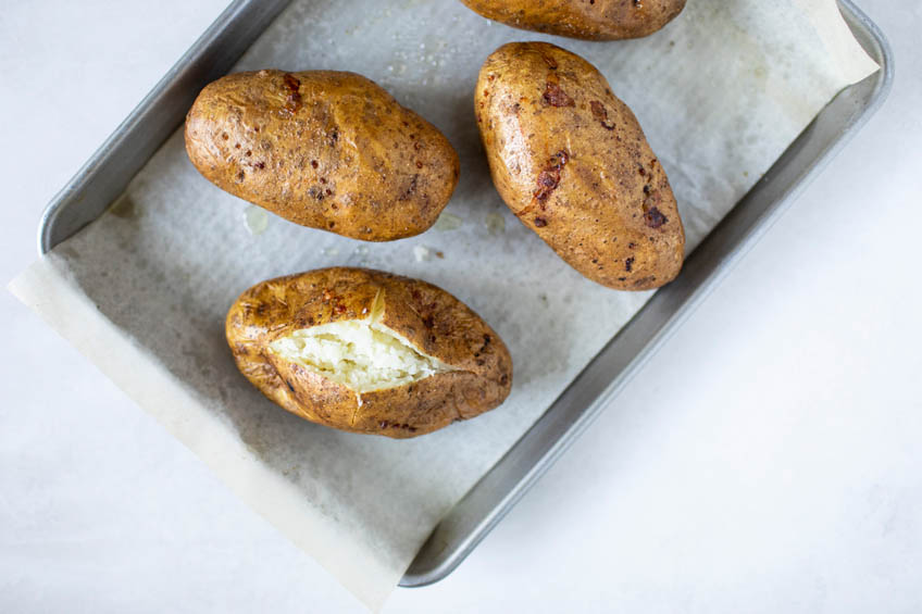 Baked potatoes on a baking tray, one is sliced open