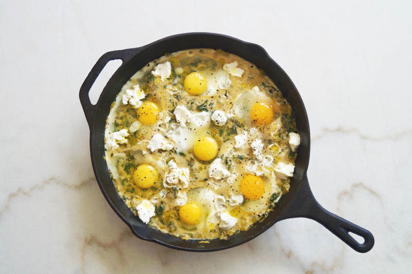 Uncooked eggs and goat cheese on a green base of veggies for creamy leek and spinach shakshuka