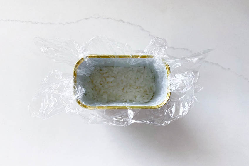 A spam can lined with plastic wrap with a layer of rice at the bottom.