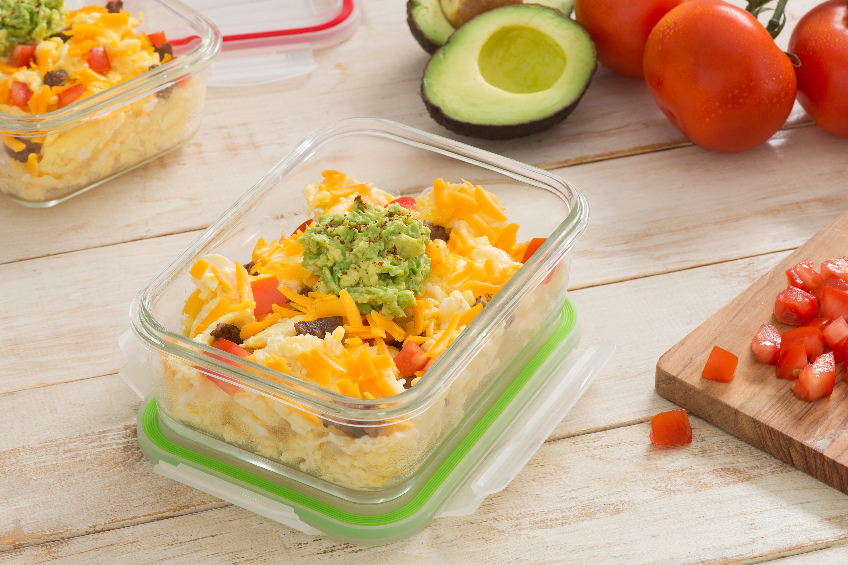A glass storage container with scrambled egg, cheese, sausage, tomato and guacamole