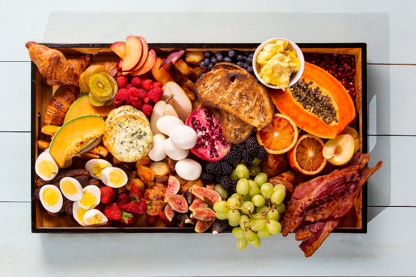 A wooden charcuterie board with eggs, bacon, fresh fruit and bread