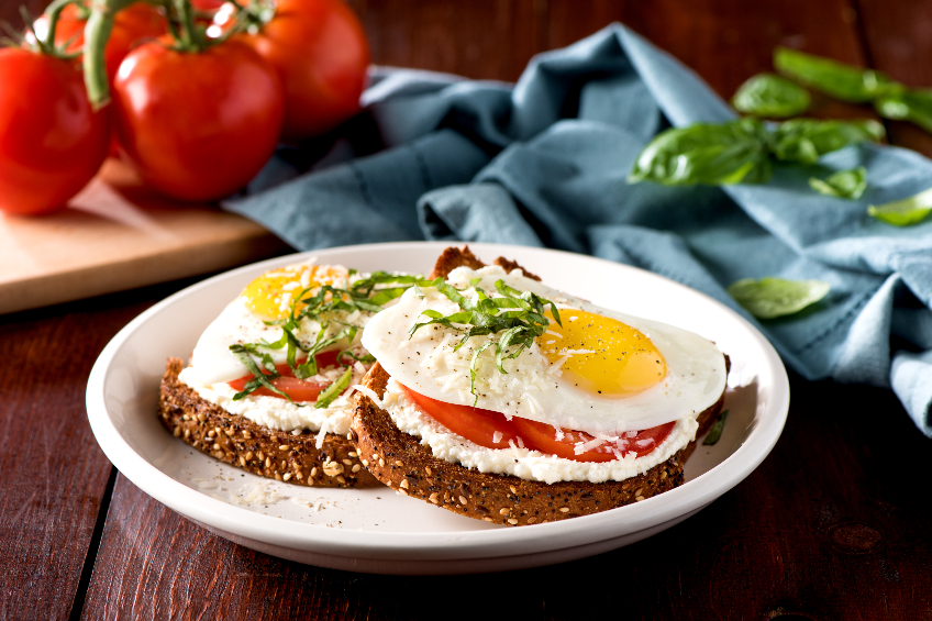 Whole grain bread topped with ricotta, sliced tomatoes and fried eggs, served on a white plate