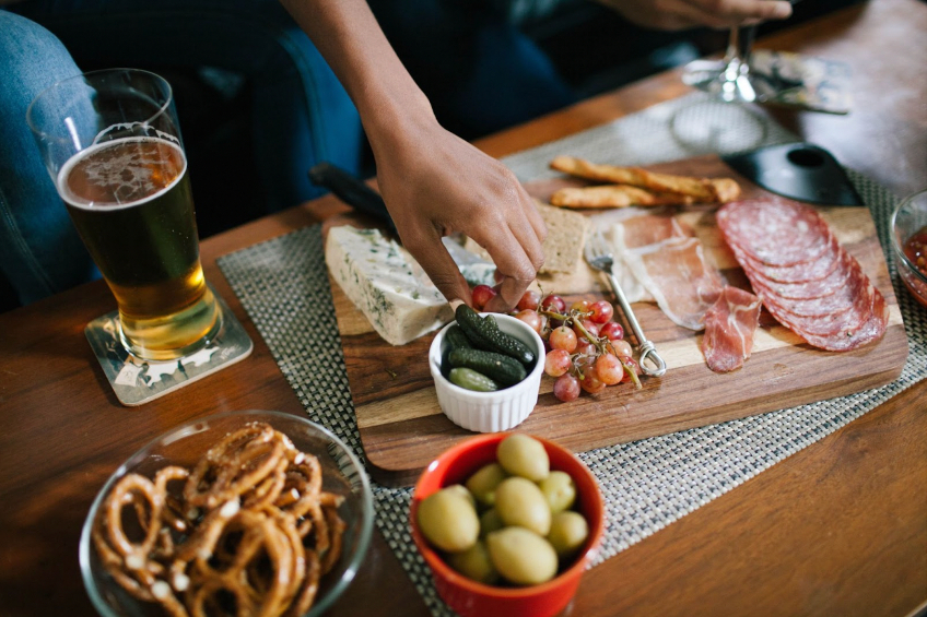 A charcuterie board with olives, meats, cheeses and pretzels is seen on a table, with a hand reaching for a bite