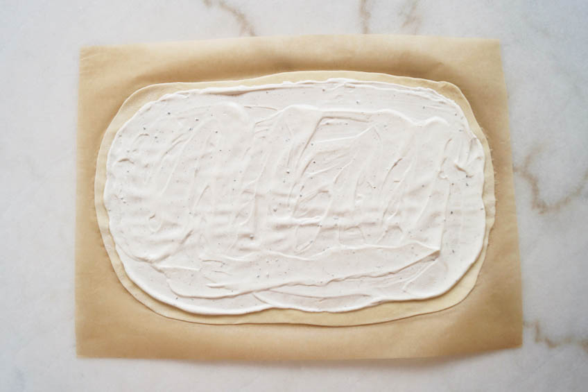 Flammkuchen dough topped with creme fraiche mixture on a sheet of parchment paper