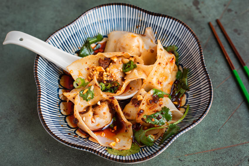 A bowl of Sichuan wontons with fresh herbs in a red chili oil