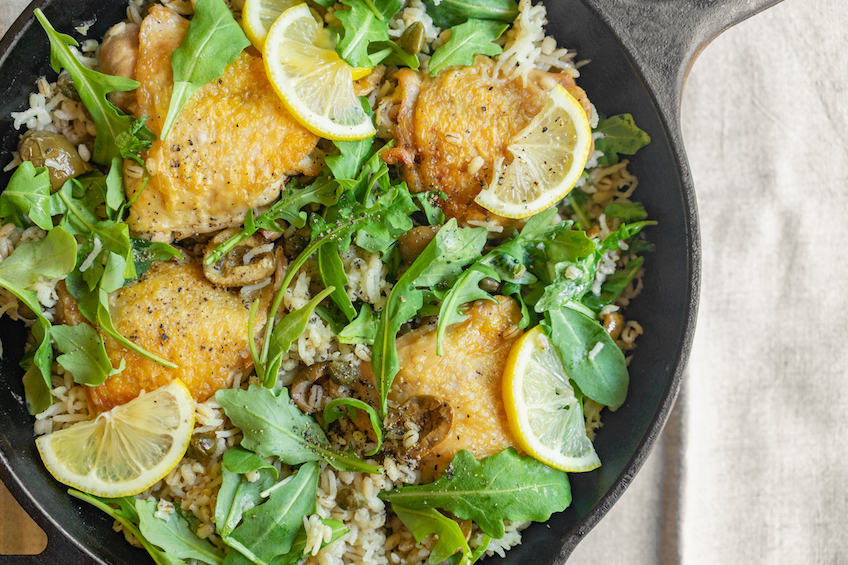 A cast iron pan with chicken thighs, rice, barley, arugula and olives