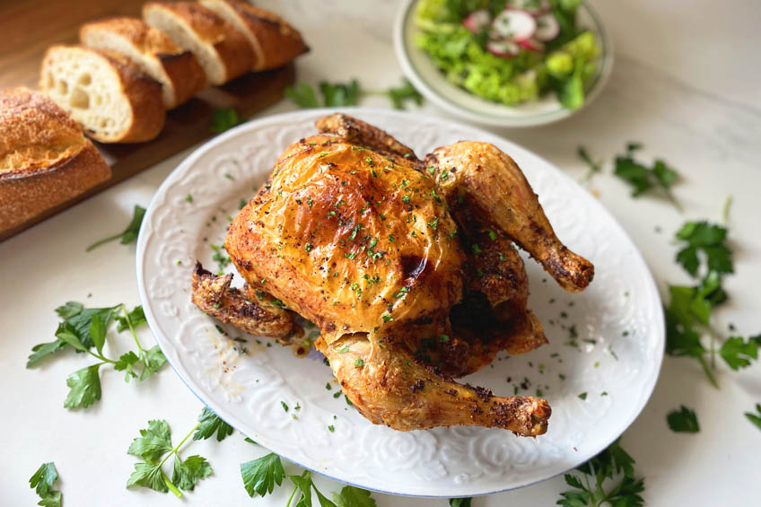 A whole roasted chicken on a serving platter, surrounded by bread and salad.