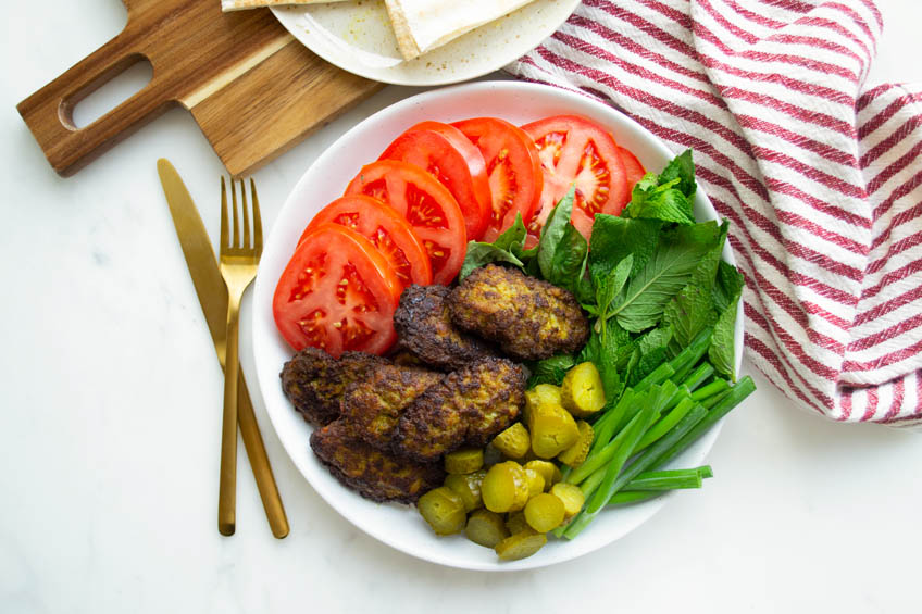 Beef kotlets on a plate with pickles, greens, and bright red tomatoes